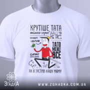 Футболка тато може все, біла, лежить на столі, фон світлий, принт з персонажем, що тримає зелений предмет, текст на футболці яскравий і кумедний.