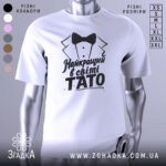 Футболка тато найкращий, біла, зображена з правого боку, фон світло-сірий, принт чорного кольору з текстом 'Найкращий в світі TATO' та малюнком смокінга.