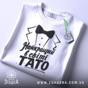 Футболка тато найкращий, біла, складена на поверхні, фон світло-сірий, принт чорного кольору з текстом 'Найкращий в світі TATO' та малюнком смокінга, з зеленим ярликом.