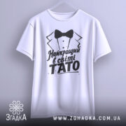 Футболка тато найкращий, біла, підвішена на вішалці, фон світло-сірий, принт чорного кольору з текстом 'Найкращий в світі TATO' та малюнком смокінга.