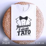 Футболка тато найкращий, біла, складена на плетеній кошику, фон світло-сірий, принт чорного кольору з текстом 'Найкращий в світі TATO' та малюнком смокінга.