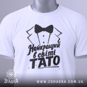 Футболка тато найкращий, біла, складена на поверхні, фон світло-сірий, принт чорного кольору з текстом 'Найкращий в світі TATO' та малюнком смокінга, з зеленим ярликом.