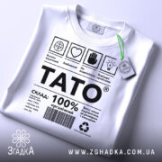 Футболка тато органічний подарунок, біла, складена на поверхні, фон світло-сірий, принт чорного кольору з текстом і символами, на комірці зелений ярлик.