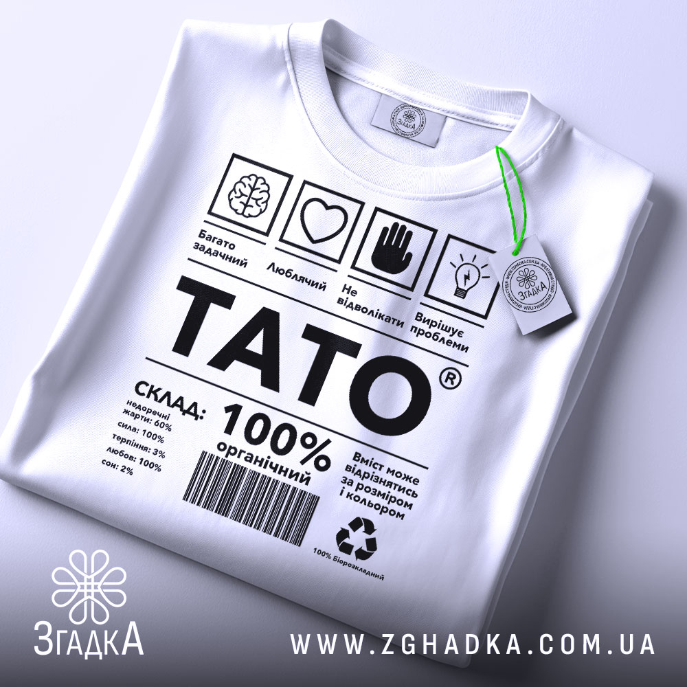 Футболка тато органічний подарунок, біла, складена на поверхні, фон світло-сірий, принт чорного кольору з текстом і символами, на комірці зелений ярлик.