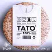 Футболка тато органічний подарунок, біла, складена на плетеній основі, фон світло-сірий, принт чорного кольору з текстом і символами.