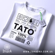 Футболка тато органічний, білого кольору, складена на плоскій поверхні, фон світло-сірий, принт чорного кольору з текстом і символами, на комірці є етикетка з зеленим шнурком.