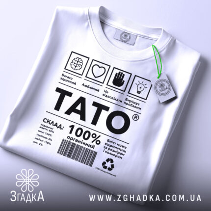 Футболка тато органічний, білого кольору, складена на плоскій поверхні, фон світло-сірий, принт чорного кольору з текстом і символами, на комірці є етикетка з зеленим шнурком.
