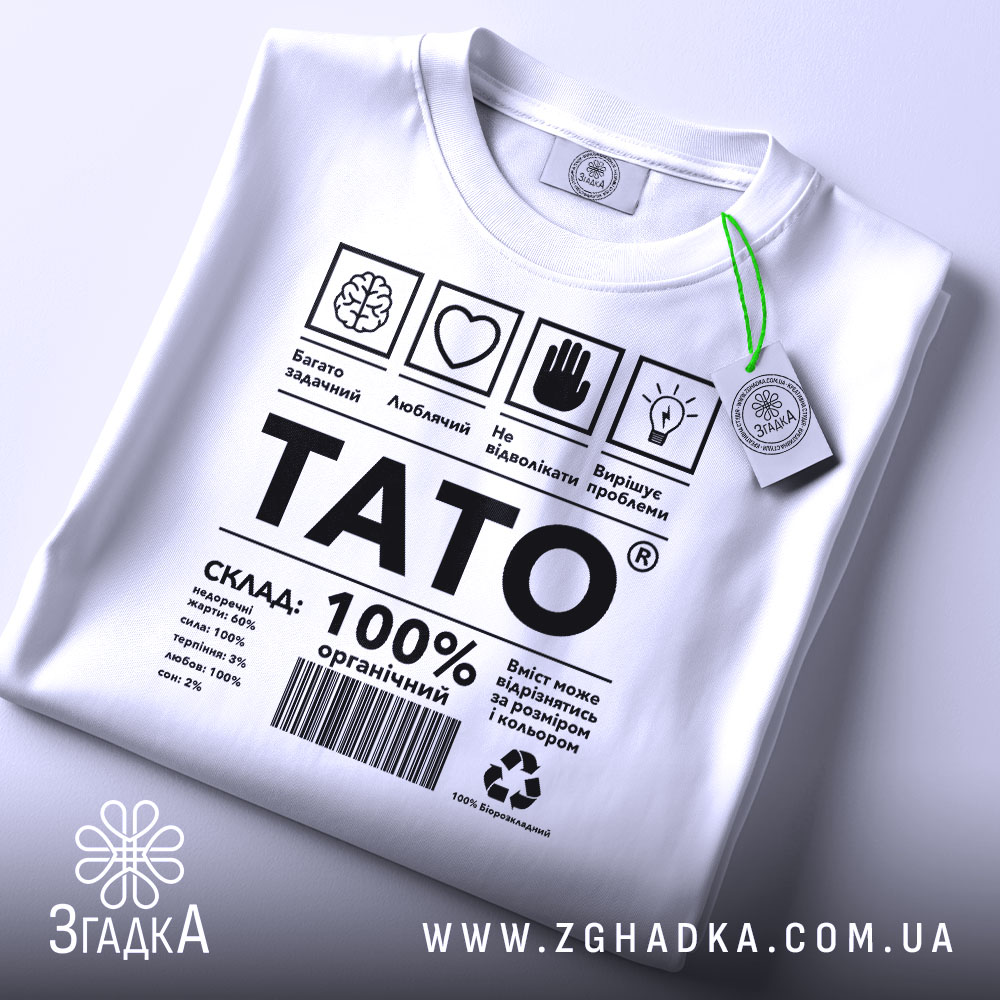 Футболка тато органічний, білого кольору, складена на плоскій поверхні, фон світло-сірий, принт чорного кольору з текстом і символами, на комірці є етикетка з зеленим шнурком.
