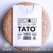 Футболка тато органічний, білого кольору, складена на плоскій поверхні з плетеним елементом, фон світлий, принт чорного кольору з текстом і символами, футболка має округлий комір.