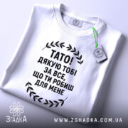 Футболка тато подяка, біла, складена на поверхні, фон світлий, принт чорний з текстом "TATO! ДЯКУЮ ТOBI ЗА ВСЕ, ЩО ТИ РОБИШ ДЛЯ МЕНЕ" та етикетка з зеленим шнурком.