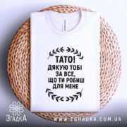 Футболка тато подяка, біла, складена на плетеній основі, фон світлий, принт чорний з текстом "TATO! ДЯКУЮ ТOBI ЗА ВСЕ, ЩО ТИ РОБИШ ДЛЯ МЕНЕ".