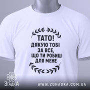 Футболка тато подяка, біла, складена на поверхні, фон світлий, принт чорний з текстом "TATO! ДЯКУЮ ТOBI ЗА ВСЕ, ЩО ТИ РОБИШ ДЛЯ МЕНЕ" та етикетка з зеленим шнурком.