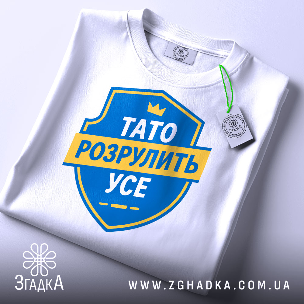 Футболка тато розрулить усе, складена на столі, з верхнім ракурсом, фон світлий, принт у вигляді синього щита з жовтою стрічкою та текстом, на комірці видна етикетка.