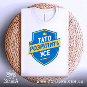 Футболка тато розрулить усе, складена на плетеній тарілці, з верхнім ракурсом, фон світлий, принт у вигляді синього щита з жовтою стрічкою та текстом.