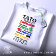 Футболка тато супергерой білого кольору, складена на поверхні, фон світлий, принт з різнокольоровими символами супергероїв та текстом, на футболці є етикетка.