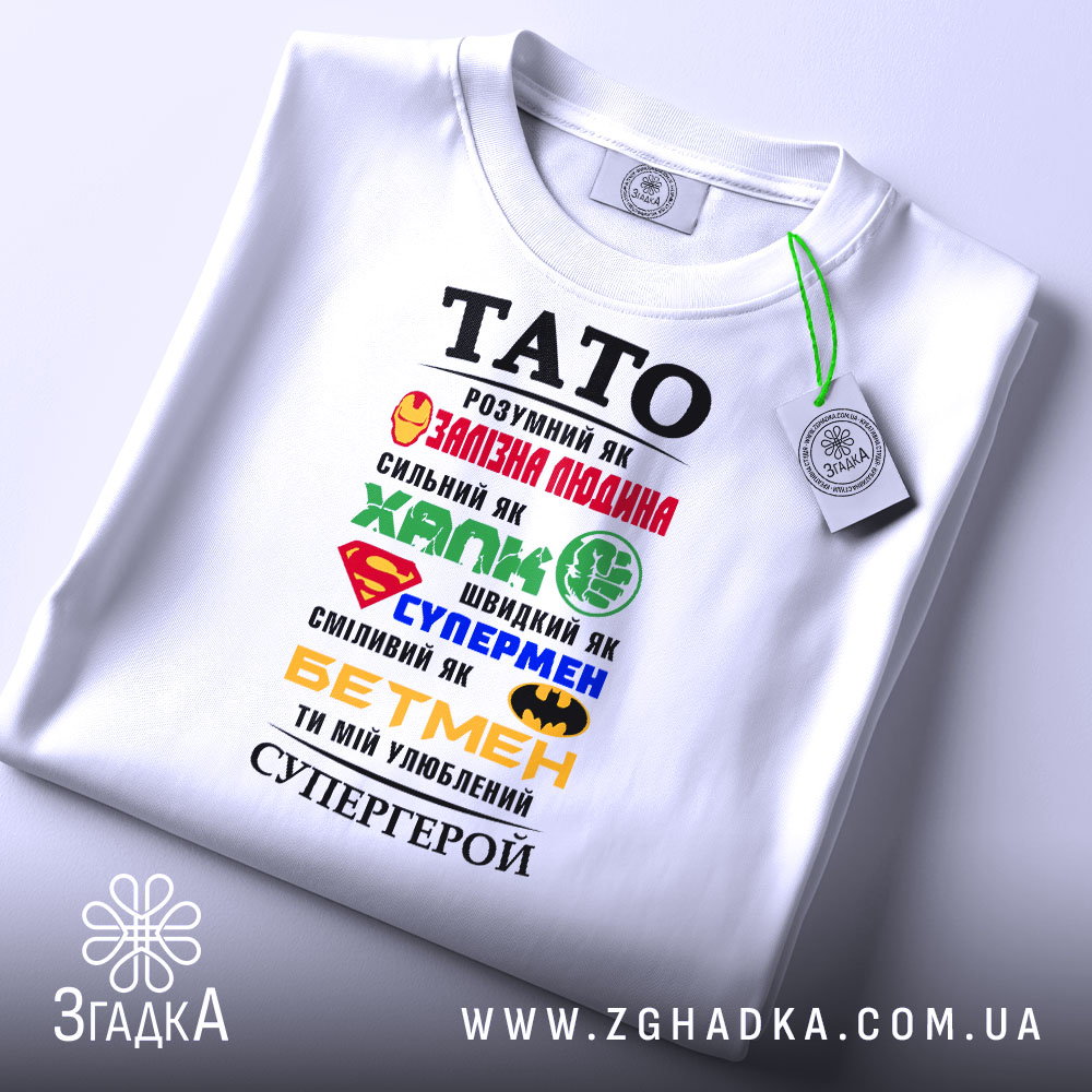 Футболка тато супергерой білого кольору, складена на поверхні, фон світлий, принт з різнокольоровими символами супергероїв та текстом, на футболці є етикетка.