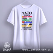 Футболка тато супергерой білого кольору, підвішена на вішалці, фон світло-сірий, принт містить різнокольорові символи супергероїв та текст, що описує тата як супергероя.