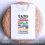 Футболка тато супергерой білого кольору, складена на плетеній основі, фон нейтральний, принт з різнокольоровими символами супергероїв та текстом, що описує тата як супергероя.