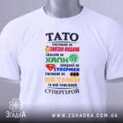 Футболка тато супергерой білого кольору, складена на поверхні, фон світлий, принт з різнокольоровими символами супергероїв та текстом, на футболці є етикетка.