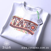 Футболка тато білого кольору, зображена в складеному вигляді, фон світло-сірий, принт з написом 'TATO' в декоративному стилі, на футболці присутня етикетка з зеленим шнурком.