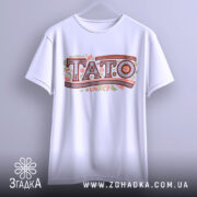 Футболка тато білого кольору, зображена з фронтального ракурсу на вішалці, фон світло-сірий, принт з написом 'TATO' в декоративному стилі з рослинними елементами.