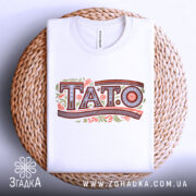 Футболка тато білого кольору, зображена в складеному вигляді на плетеній основі, фон світло-сірий, принт з написом 'TATO' в декоративному стилі.