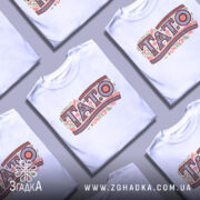 Футболка тато білого кольору, зображена з фронтального ракурсу на манекені, фон світло-сірий, принт з написом 'TATO' в декоративному стилі з рослинними елементами.