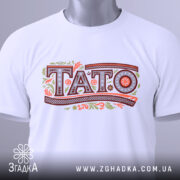 Футболка тато білого кольору, зображена в складеному вигляді, фон світло-сірий, принт з написом 'TATO' в декоративному стилі, на футболці присутня етикетка з зеленим шнурком.