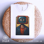 Футболка телевізор око, біла, зображена зверху на плетеній кошику, фон світло-сірий, принт з телевізором і оком на голові персонажа, видима етикетка.