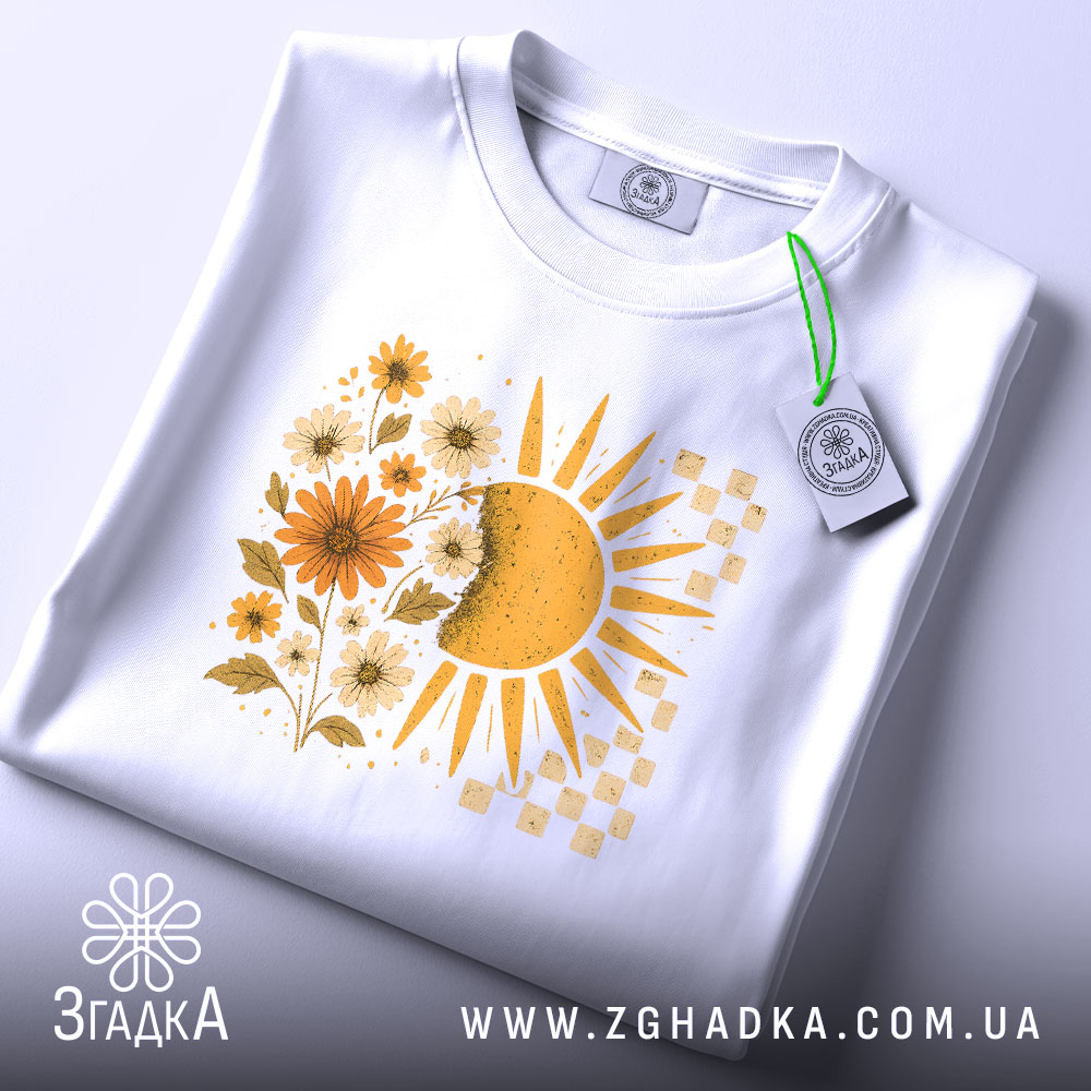 Футболка тепло життя. Біла футболка, складена на столі, з принтом сонця і квітів жовтого кольору. На футболці видно етикетку з зеленим шнурком, фон світло-сірий.
