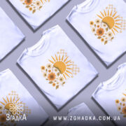 Футболка тепло життя. Біла футболка з круглим вирізом, зображена з фронтального ракурсу на манекені. На футболці яскравий принт з сонцем і квітами жовтого кольору, фон світло-сірий.