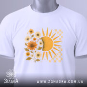 Футболка тепло життя. Біла футболка, складена на столі, з принтом сонця і квітів жовтого кольору. На футболці видно етикетку з зеленим шнурком, фон світло-сірий.