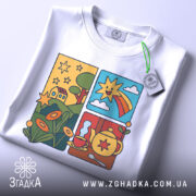 Футболка теплий ранок. Біла футболка, складена на поверхні, принт яскравий, з елементами природи, сонце та чайник, на футболці є етикетка з зеленим шнурком.