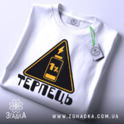 Футболка терпець білого кольору, складена на поверхні, ракурс зверху, фон світло-сірий, принт у вигляді жовтого трикутника з чорним написом 'ТЕРПЕЦЬ' та зображенням батареї з 1% заряду, на комірці є етикетка.