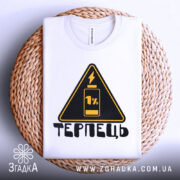 Футболка терпець білого кольору, складена на плетеній кошику, ракурс зверху, фон світло-сірий, принт у вигляді жовтого трикутника з чорним написом 'ТЕРПЕЦЬ' та зображенням батареї з 1% заряду.