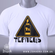 Футболка терпець білого кольору, складена на поверхні, ракурс зверху, фон світло-сірий, принт у вигляді жовтого трикутника з чорним написом 'ТЕРПЕЦЬ' та зображенням батареї з 1% заряду, на комірці є етикетка.