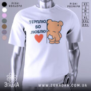 Футболка терплю люблю, білого кольору, зображена на манекені з легким боковим ракурсом, фон світло-сірий, принт з ведмедиком, що тримається за стіну, з написом 'ТЕРПЛЮ, БО ЛЮБЛЮ!' і червоним сердечком.