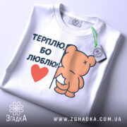 Футболка терплю люблю, білого кольору, складена на поверхні, з верхнього ракурсу, фон світло-сірий, принт з ведмедиком, що тримається за стіну, з написом 'ТЕРПЛЮ, БО ЛЮБЛЮ!' і червоним сердечком, на комірці видна етикетка.