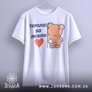 Футболка терплю люблю, білого кольору, підвішена на вішалці, фон світло-сірий, принт з ведмедиком, що тримається за стіну, з написом 'ТЕРПЛЮ, БО ЛЮБЛЮ!' і червоним сердечком.