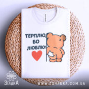 Футболка терплю люблю, білого кольору, складена на плетеній кошику, фон білий, принт з ведмедиком, що тримається за стіну, з написом 'ТЕРПЛЮ, БО ЛЮБЛЮ!' і червоним сердечком, видна етикетка на комірці.