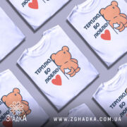 Футболка терплю люблю, білого кольору, зображена на манекені з легким боковим ракурсом, фон світло-сірий, принт з ведмедиком, що тримається за стіну, з написом 'ТЕРПЛЮ, БО ЛЮБЛЮ!' і червоним сердечком.