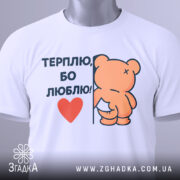 Футболка терплю люблю, білого кольору, складена на поверхні, з верхнього ракурсу, фон світло-сірий, принт з ведмедиком, що тримається за стіну, з написом 'ТЕРПЛЮ, БО ЛЮБЛЮ!' і червоним сердечком, на комірці видна етикетка.