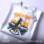 Футболка this is fine, біла, складена на столі, фон світло-сірий, принт з собакою на фоні двох автомобілів, яскраві кольори: оранжевий, чорний, жовтий, з етикеткою на шнурку.