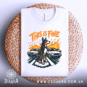 Футболка this is fine, біла, складена на плетеному кошику, фон світло-сірий, принт з собакою на фоні двох автомобілів, яскраві кольори: оранжевий, чорний, жовтий.