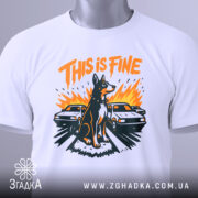 Футболка this is fine, біла, складена на столі, фон світло-сірий, принт з собакою на фоні двох автомобілів, яскраві кольори: оранжевий, чорний, жовтий, з етикеткою на шнурку.