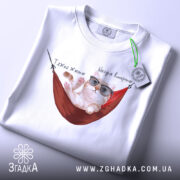 Футболка тяжко жити, біла, складена на столі, фон світло-сірий, принт з котом у окулярах, що лежить у гамаку, з написом 'Тяжко жити' і 'Шкода вмерти', на ярлику зелена стрічка.