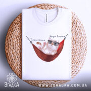 Футболка тяжко жити, біла, складена на плетеному кошику, фон світло-сірий, принт з котом у окулярах, що лежить у гамаку, з написом 'Тяжко жити' і 'Шкода вмерти'.