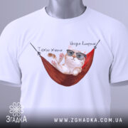 Футболка тяжко жити, біла, складена на столі, фон світло-сірий, принт з котом у окулярах, що лежить у гамаку, з написом 'Тяжко жити' і 'Шкода вмерти', на ярлику зелена стрічка.