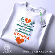 Футболка токсік бусінка, біла, зображена зверху, фон світлий, принт з текстом у зеленому та оранжевому кольорах, з серцем та рослинними елементами, на комірці є етикетка.