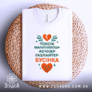 Футболка токсік бусінка, біла, зображена на плетеній основі, фон світлий, принт з текстом у зеленому та оранжевому кольорах, з серцем та рослинними елементами, видна етикетка на комірці.